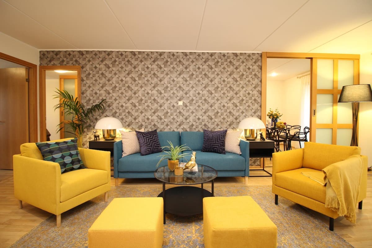 Una sala de estar con un sofá azul, dos sillones amarillos, una mesita de centro y una pared con diseño.