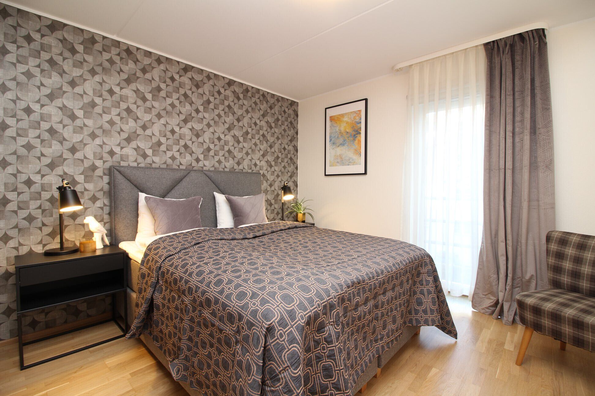 A bedroom with a patterned bedspread, a headboard, bedside lamps, a framed artwork, and a plaid chair.