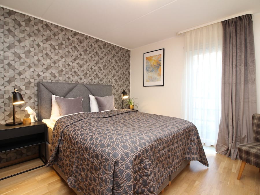 A bedroom with a patterned bedspread, a headboard, bedside lamps, a framed artwork, and a plaid chair.