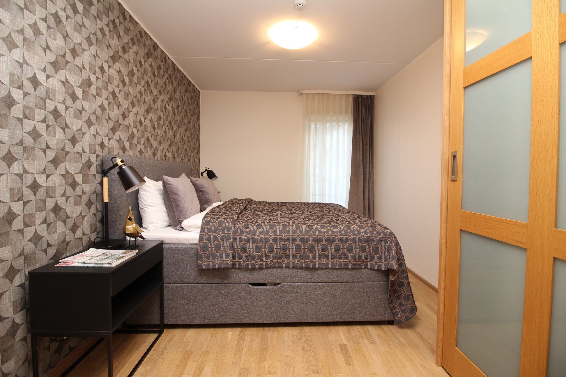 Une chambre à coucher avec un papier peint à motifs, un lit recouvert d’une couvre-lit à motifs, une table de chevet avec une lampe et une porte en bois.