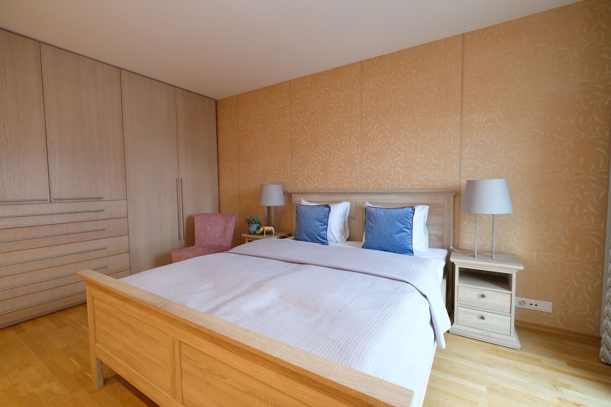 Een slaapkamer met een houten bed, twee nachtkastjes met lampen, een bijzettafeltje en een roze stoel.