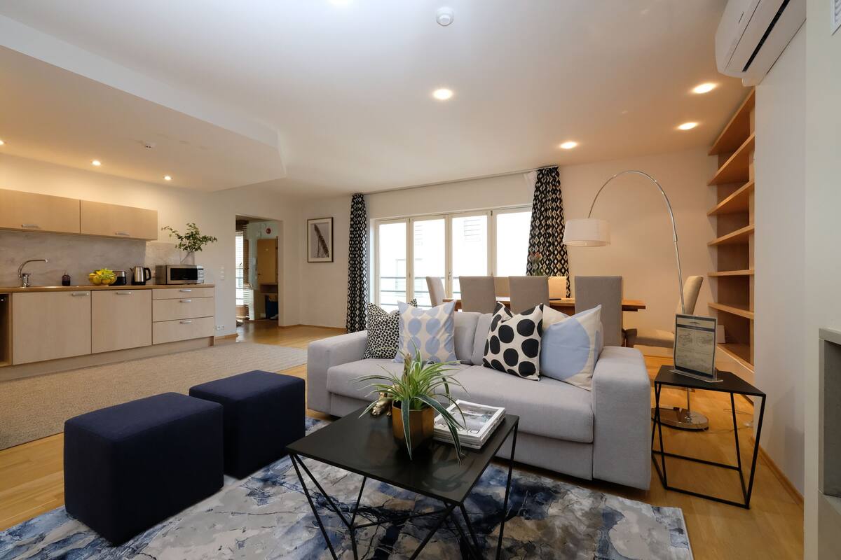 Una sala de estar moderna con sofá, mesa de centro y otomanas. Cuenta con una zona de cocina con armarios y un lavabo.