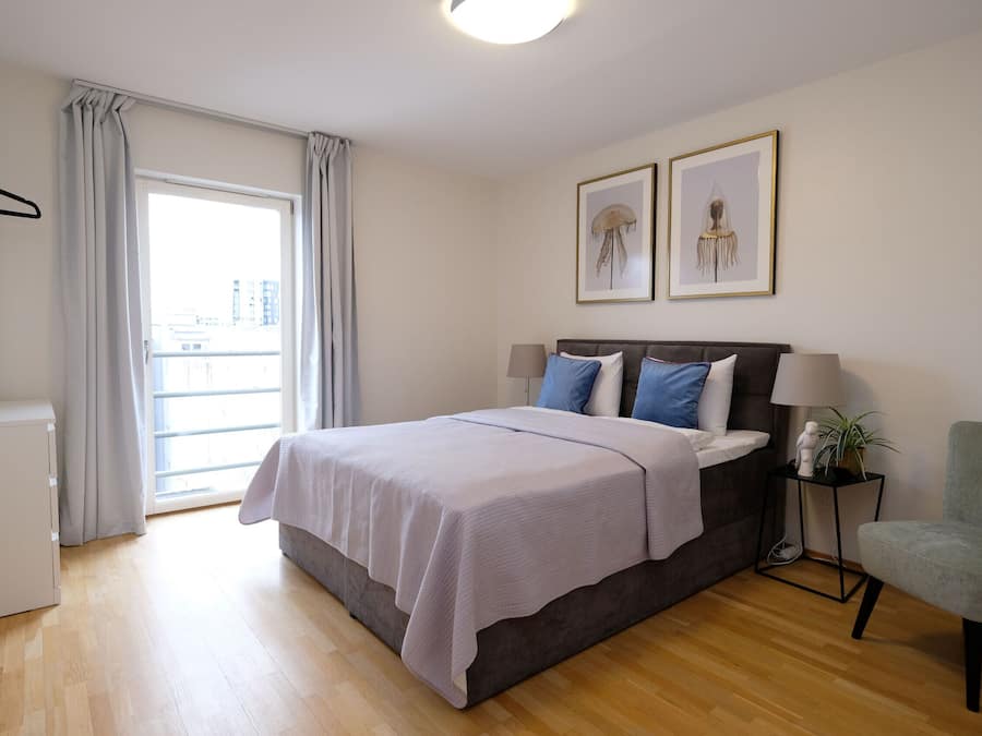 Un dormitorio con una cama, una silla, una mesita de noche con lámpara y dos cuadros en la pared.