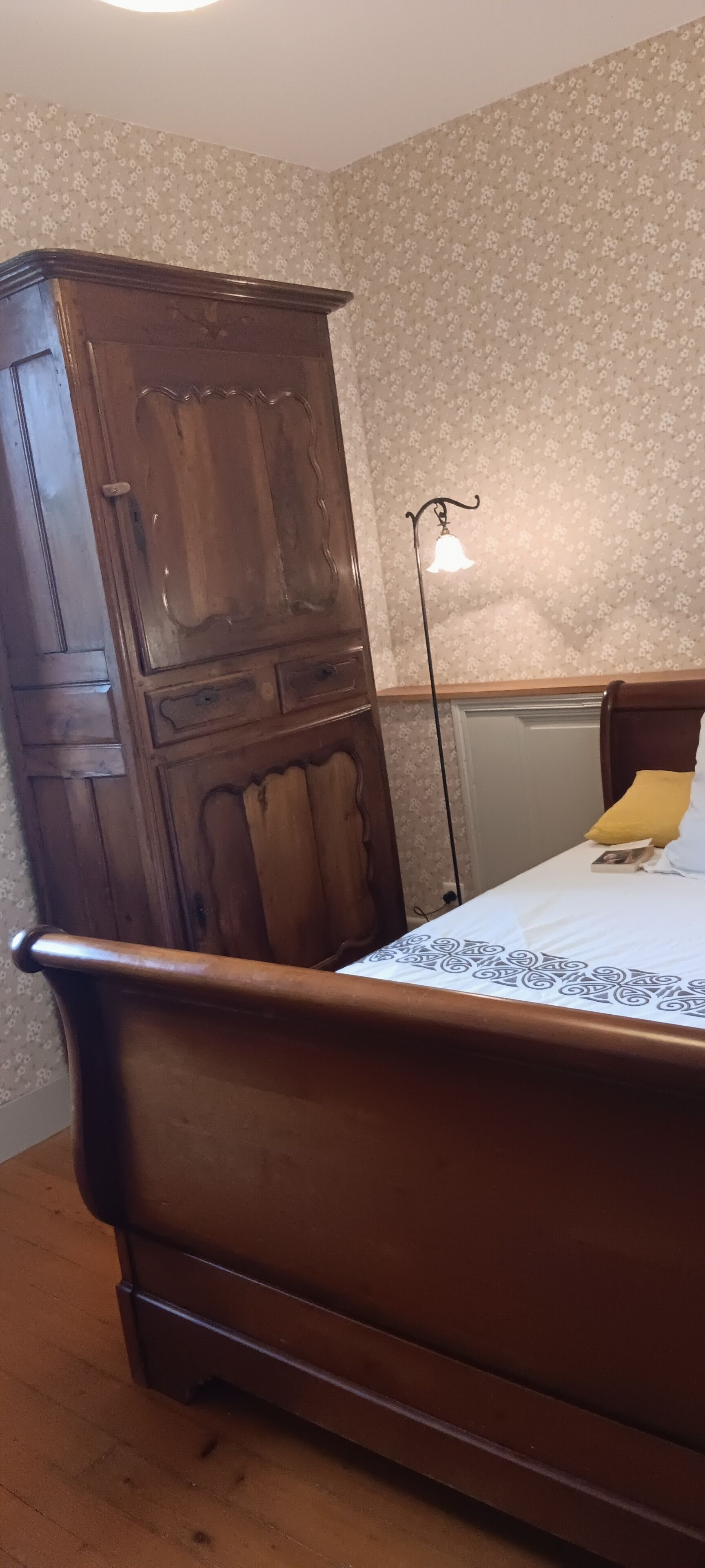 A wooden bed with a white sheet and a yellow pillow, a wooden wardrobe, and a wall with floral wallpaper.