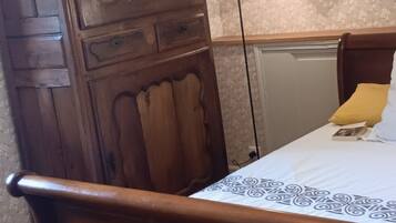 A wooden bed with a white sheet and a yellow pillow, a wooden wardrobe, and a wall with floral wallpaper.