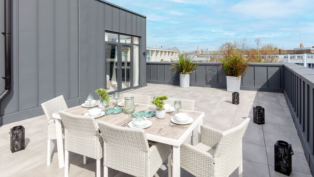 A rooftop terrace with a dining table set for four, surrounded by wicker chairs. There are potted plants and decorative lanterns on the terrace.