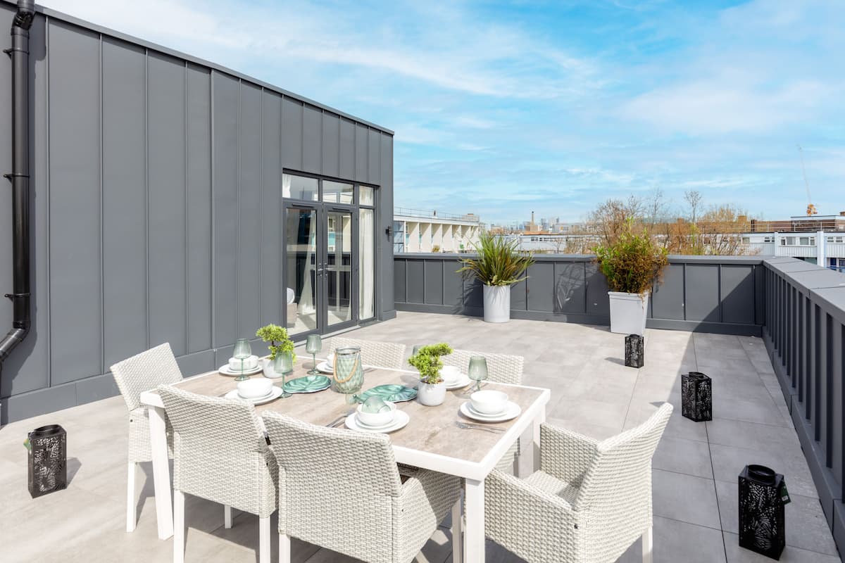 Een dakterras met een eettafel voor vier personen, omringd door rieten stoelen. Er staan potplanten en decoratieve lantaarns op het terras.