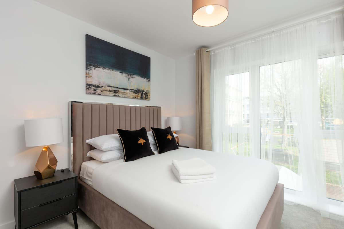 Een moderne hotelkamer met een groot bed, een nachtkastje met een lamp, een schilderij aan de muur en uitzicht op groen buiten het raam.