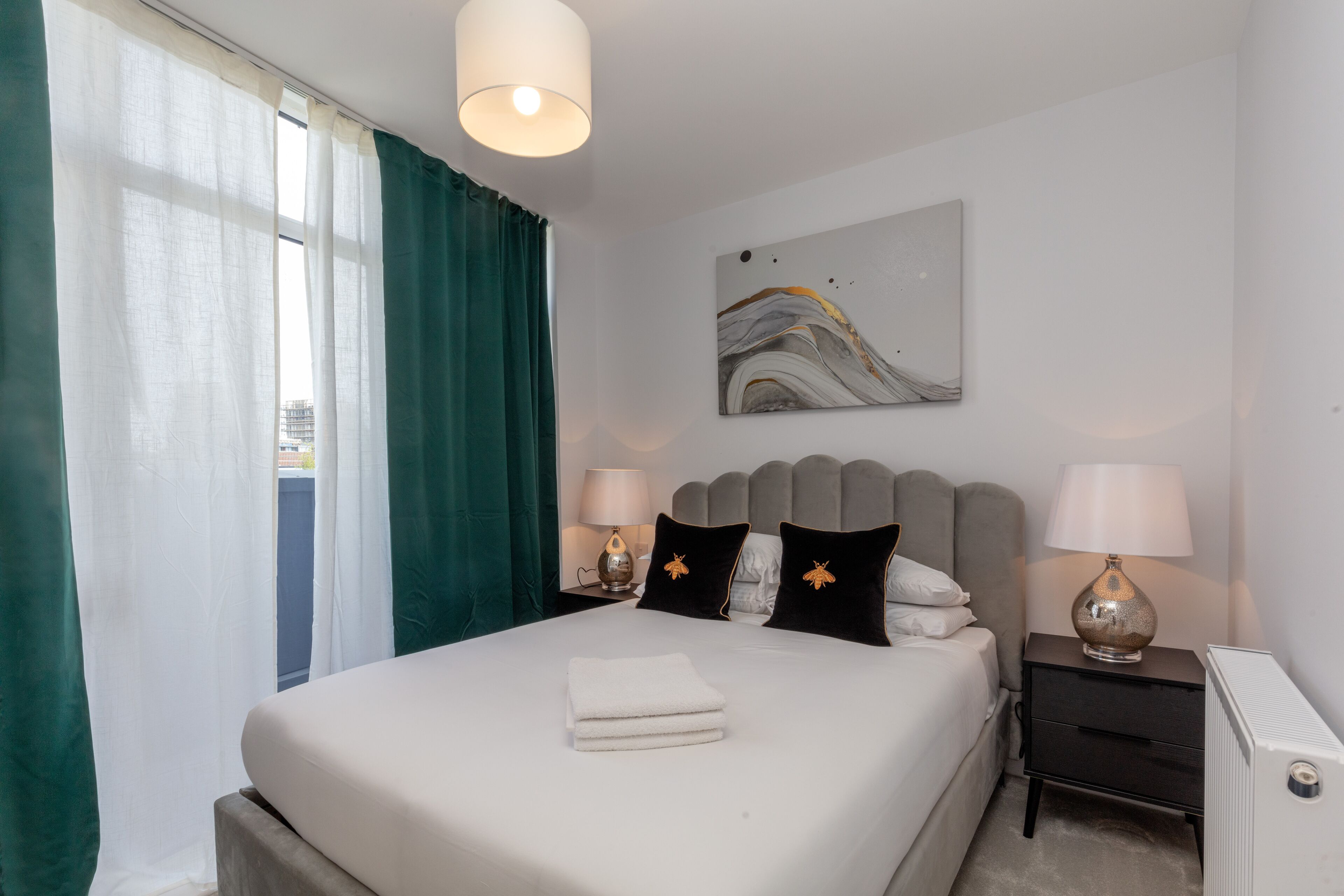 A neatly made bed with white linens, two bedside tables with lamps, a wall-mounted abstract painting, and a window with sheer curtains.