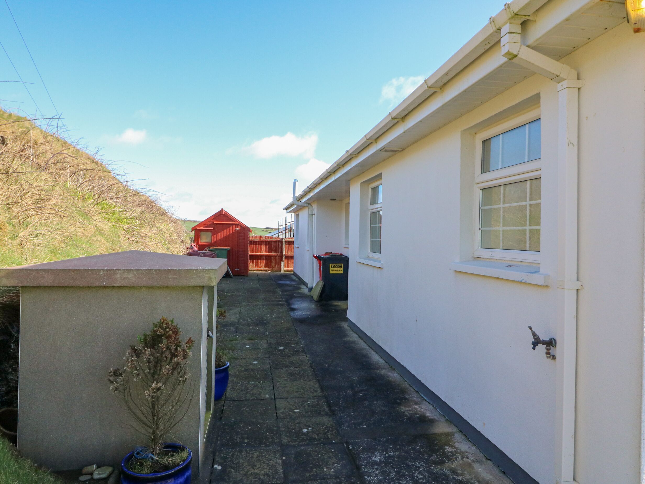 A narrow pathway leading to a white building with a window and a door. There is a small red structure in the distance, a blue trash bin, and a potted plant on the left side.