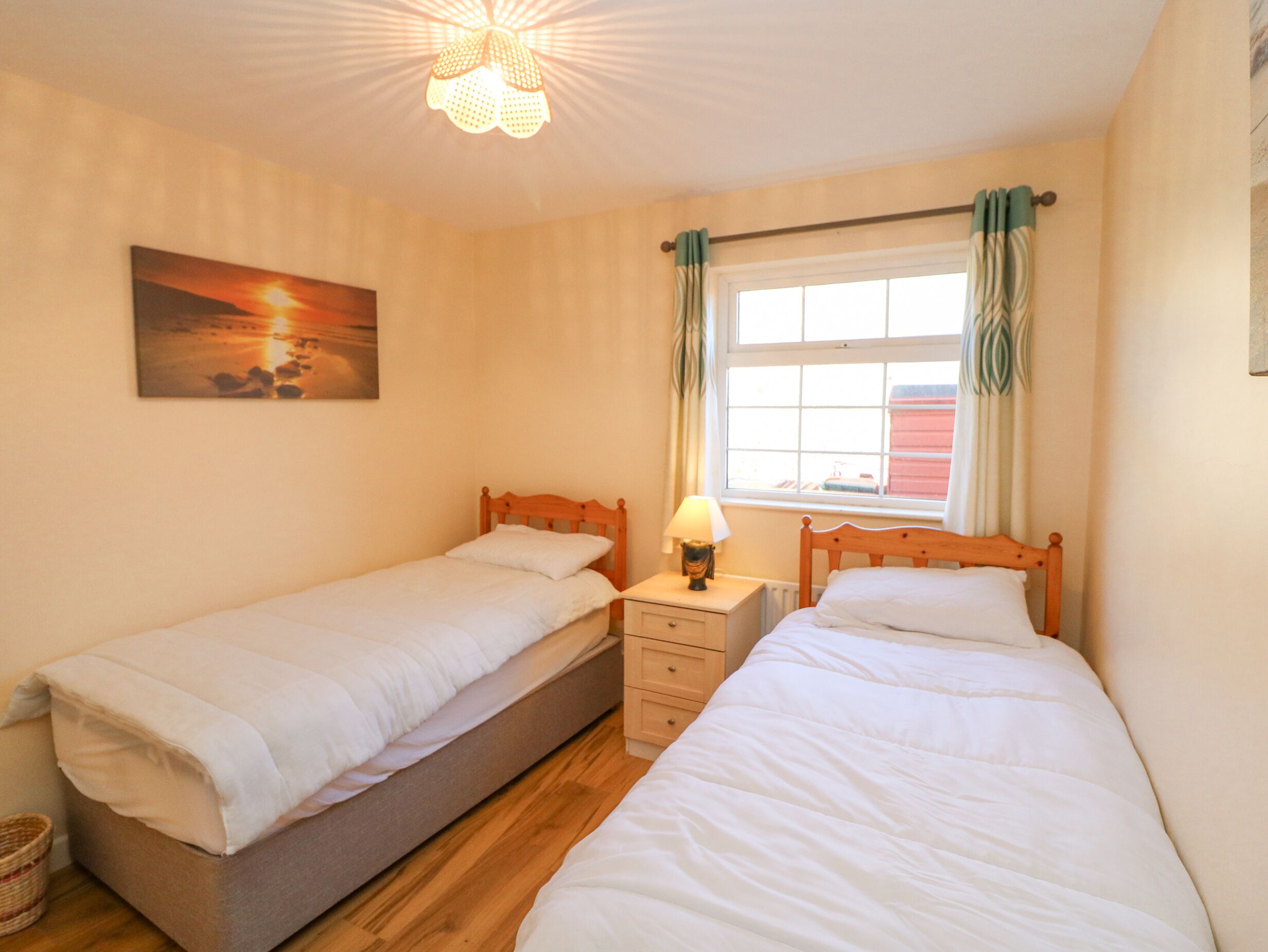 A bedroom with two single beds, a nightstand with a lamp, a window with curtains, and a painting on the wall.