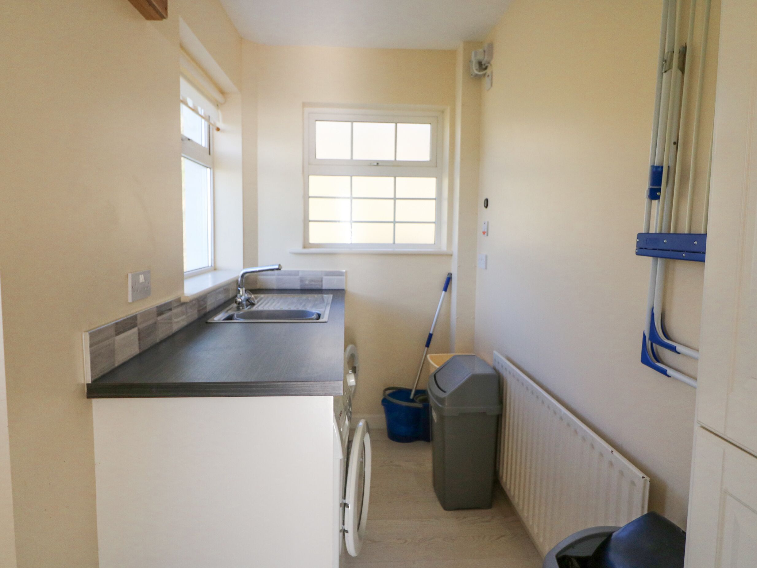 A small kitchen with a sink, a window, a mop, and a trash bin.
