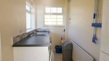 A small kitchen with a sink, a window, a mop, and a trash bin.