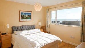 A bedroom with a large bed, a window with a view of the sea, a painting on the wall, and a lamp on each side of the bed. - Sea's The Dream (Clonakilty)