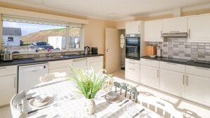 A modern kitchen with white cabinets, a dining table set for four, and a view of a car and hills outside. - Sea's The Dream (Clonakilty)