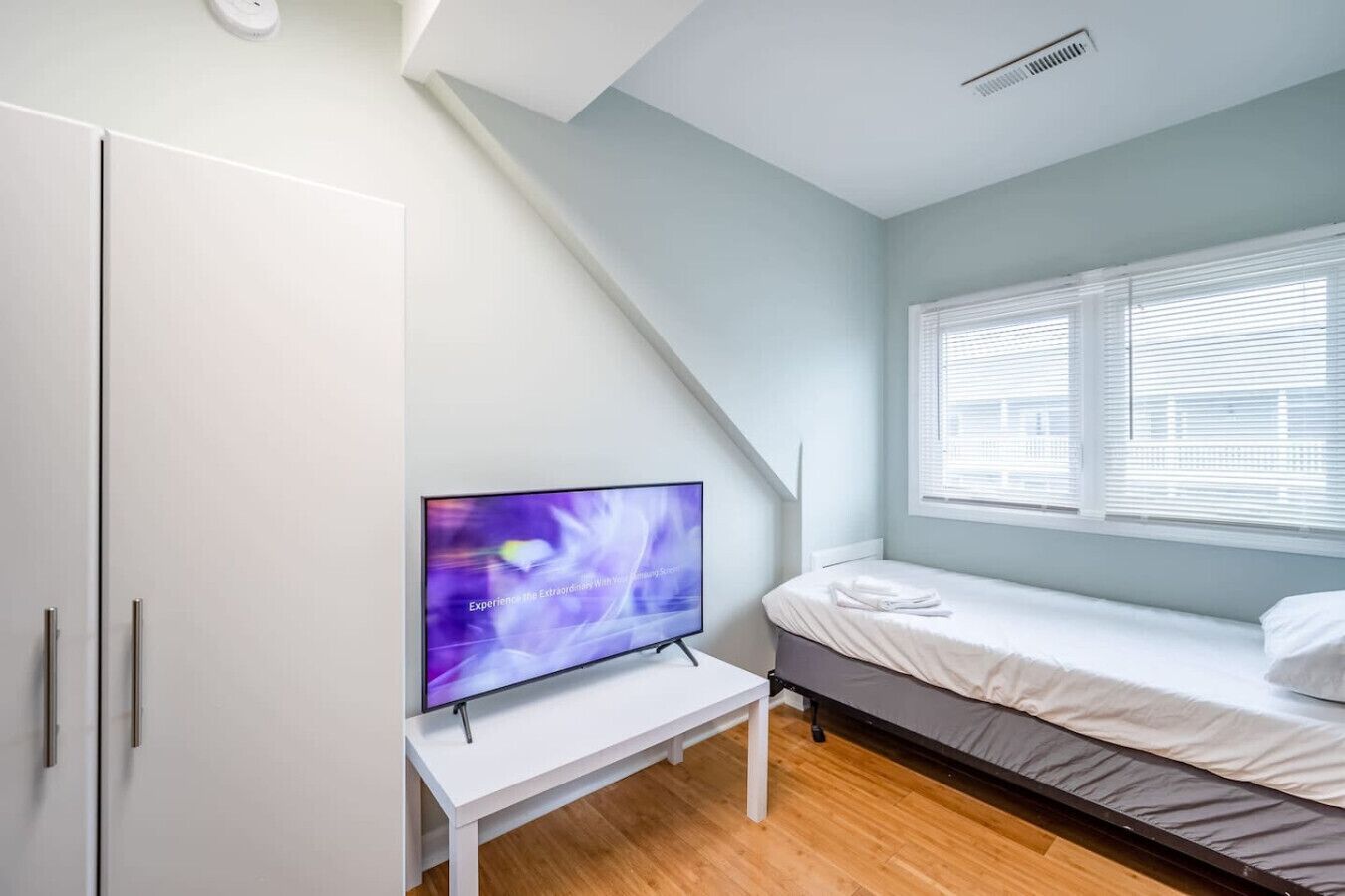 A bedroom with a bed, a TV on a white desk, and a window with blinds.