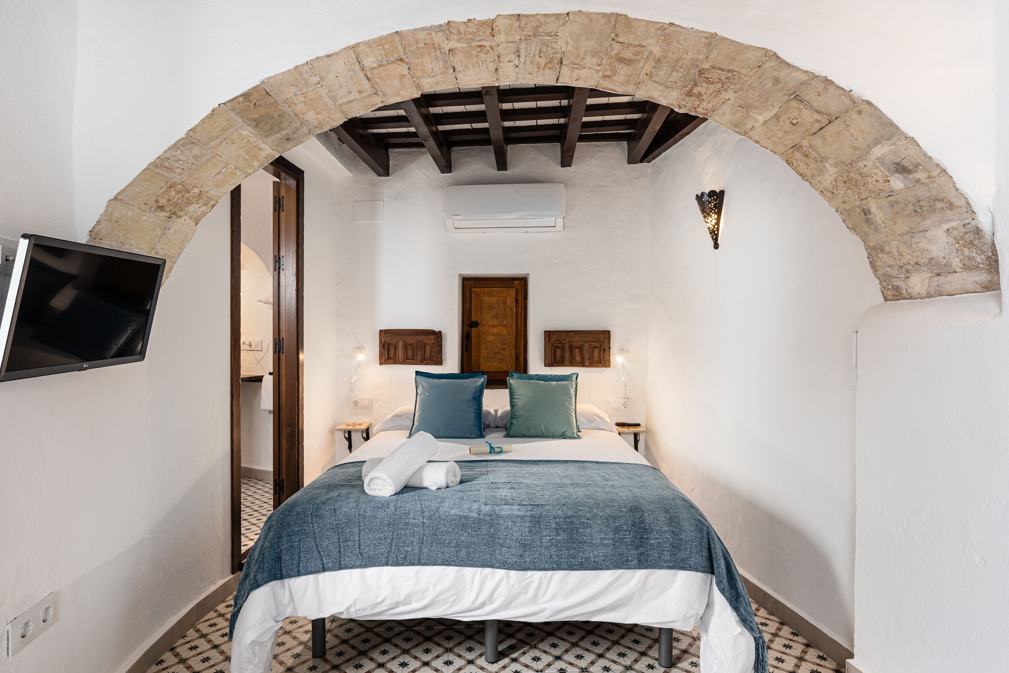 Un dormitorio con un arco de piedra, una cama con colcha azul y blanca, dos mesitas de noche con lámparas y un televisor de pantalla plana montado en la pared.