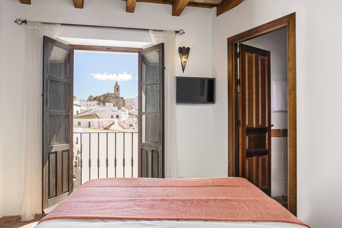 Une chambre avec un lit, une porte ouverte donnant sur un balcon, un téléviseur fixé au mur et une vue sur des immeubles et une tour à l’extérieur.