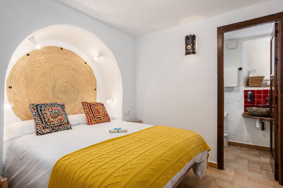 Une chambre à coucher avec un lit, une tête de lit dotée de lampes, un miroir et une salle de bain visible à travers une porte ouverte.