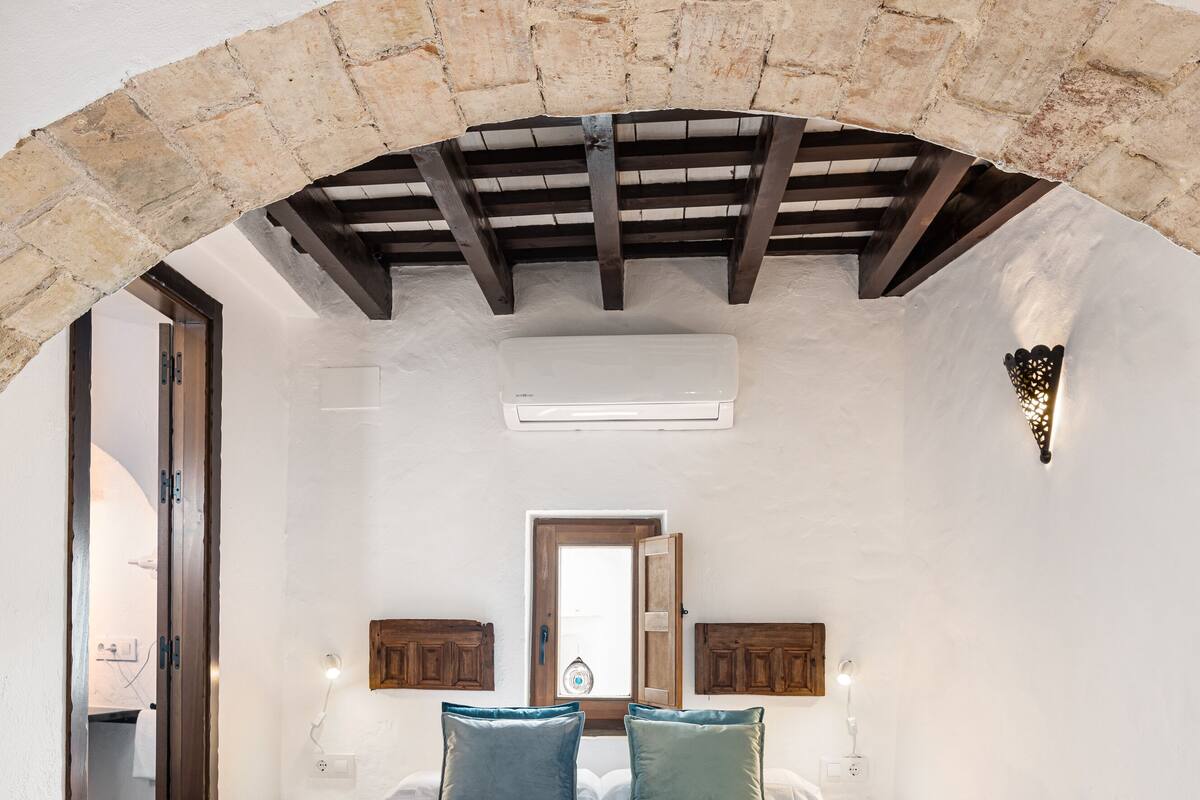Une chambre avec un grand lit, une arche en pierre, des poutres apparentes, un plafond blanc et un sol carrelé.