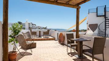 Un patio exterior con mobiliario de mimbre, una planta en maceta y vista al mar y edificios blancos al fondo.