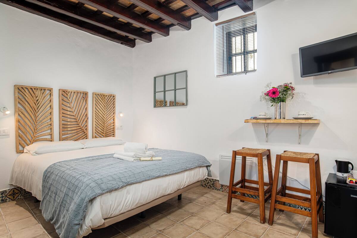 Une chambre à coucher avec un lit, des panneaux en bois formant la tête de lit, une table de chevet avec un vase de fleurs, un téléviseur fixé au mur et une fenêtre avec des stores.