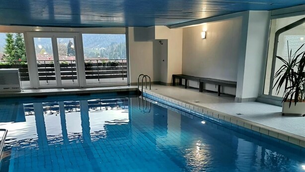 An indoor swimming pool with clear blue water, tiled floor, and a view of greenery outside. - Wohnung 06 (Berchtesgaden)
