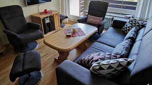 A modern living room with a sofa, two armchairs, a wooden coffee table, a TV, and a window with a view of trees. - Wohnung 06 (Berchtesgaden)