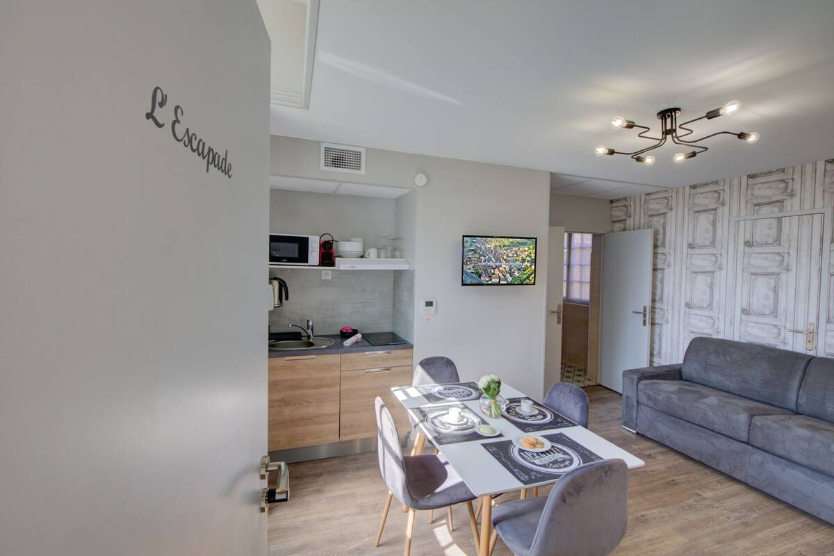 Een moderne woonkamer met een grijze bank, een eettafel gedekt voor twee personen en een keuken met een magnetron en spoelbak.