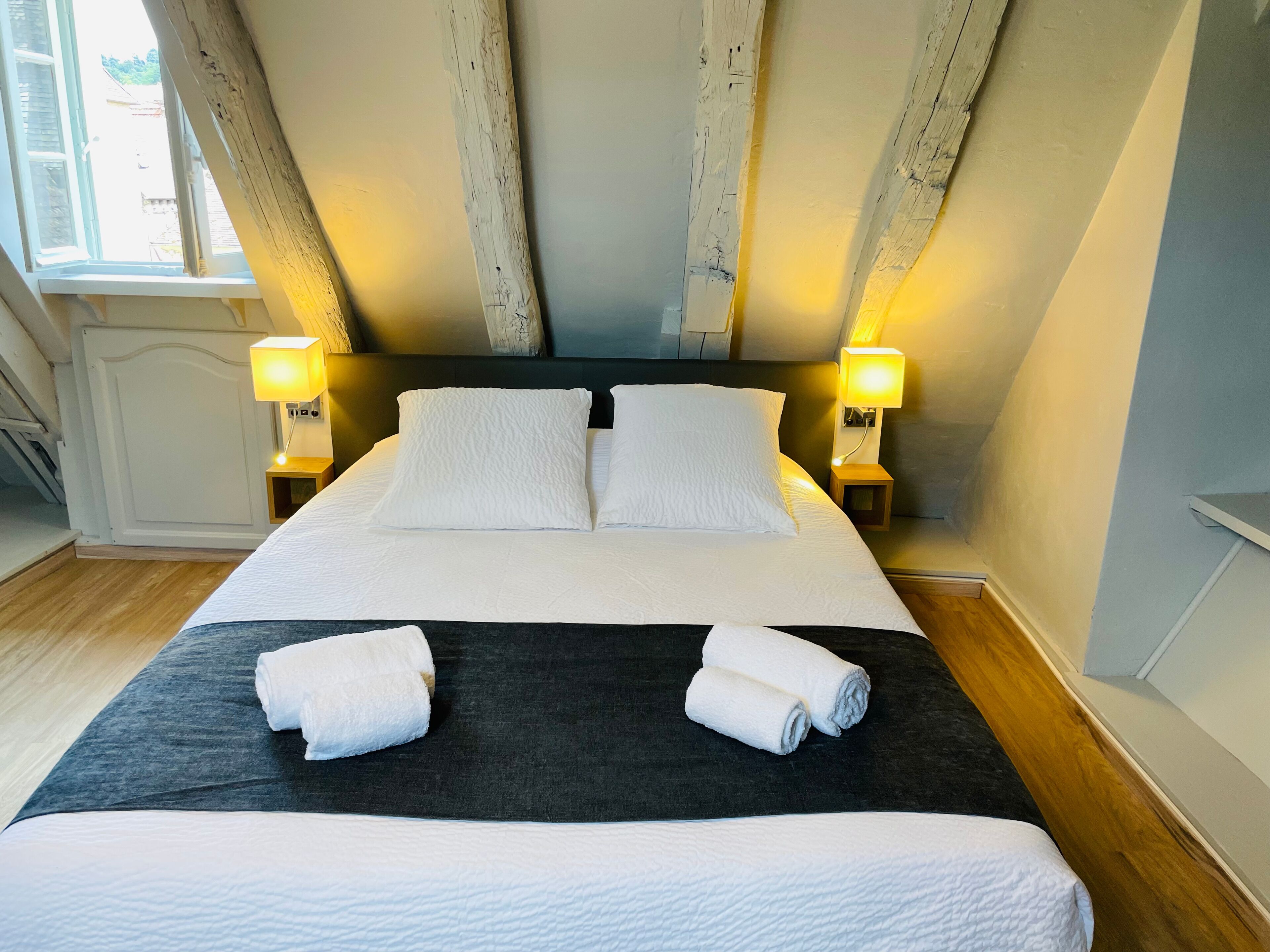 A neatly made bed with white linens and two rolled towels, two bedside lamps, and a window with a view of the outside.
