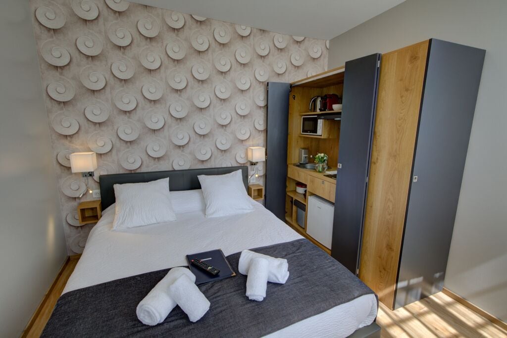 Een netjes opgemaakt bed met witte lakens, een grijze deken en twee opgerolde handdoeken. Er staat een nachtkastje met een lamp en een afstandsbediening. De kamer heeft een modern design met een behang met patroon en een houten garderobe.