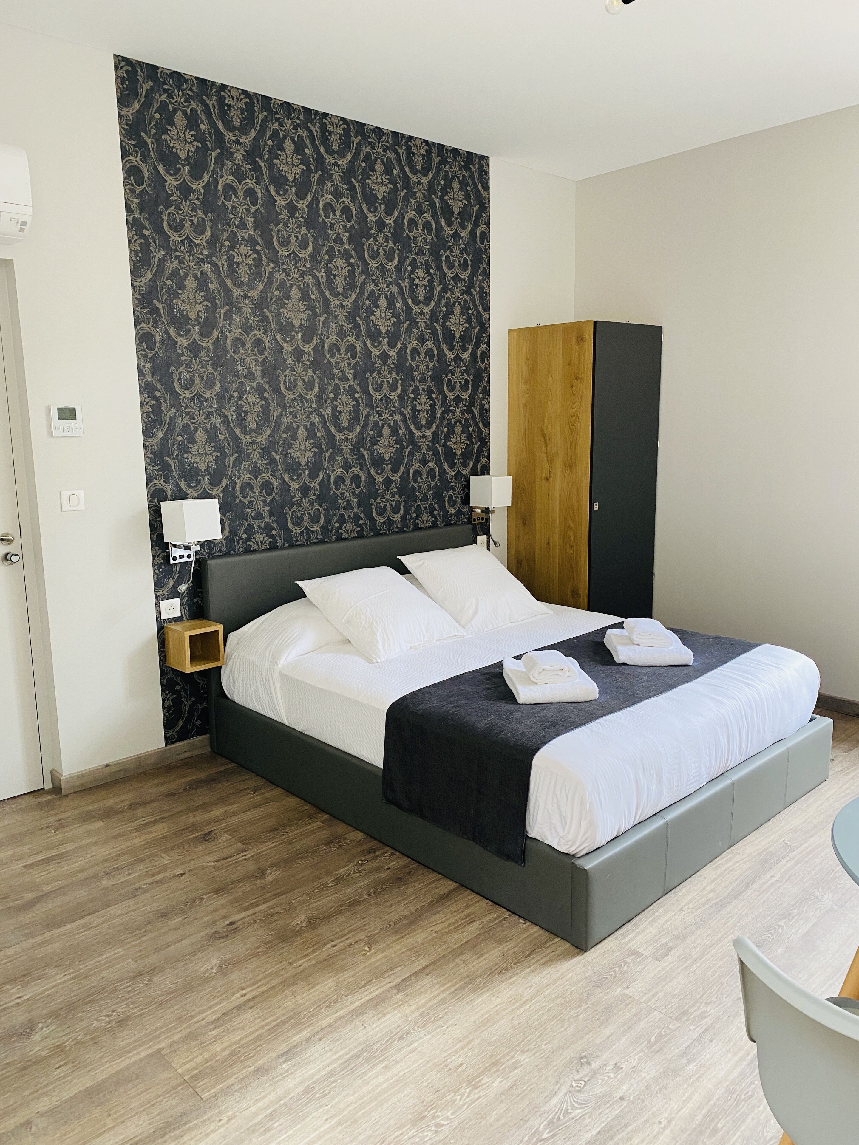 A modern hotel room with a large bed, a wooden wardrobe, and a small bedside table with a lamp.
