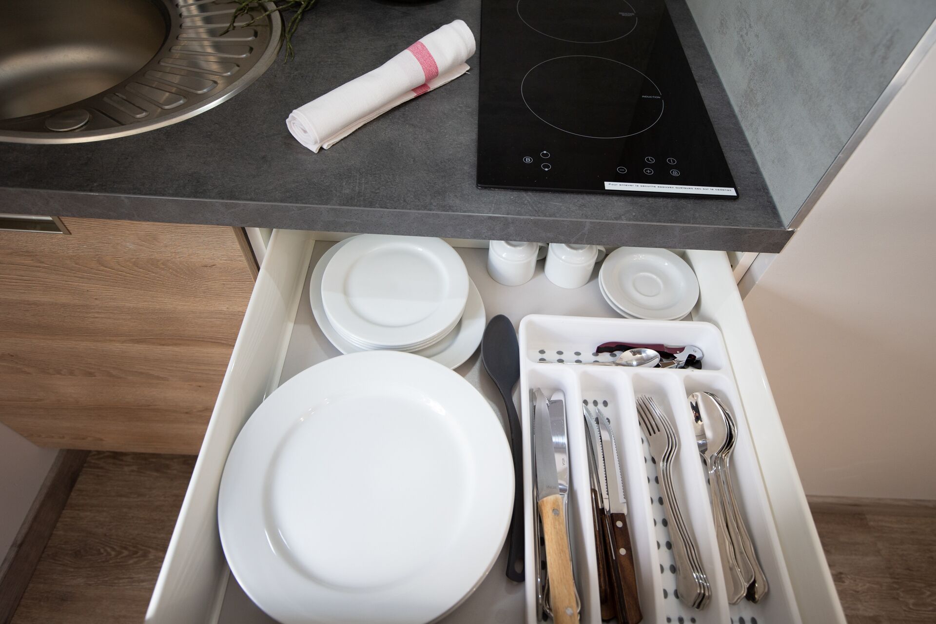 A kitchen with a built-in stove, a sink, and a drawer with neatly arranged dishes and utensils.