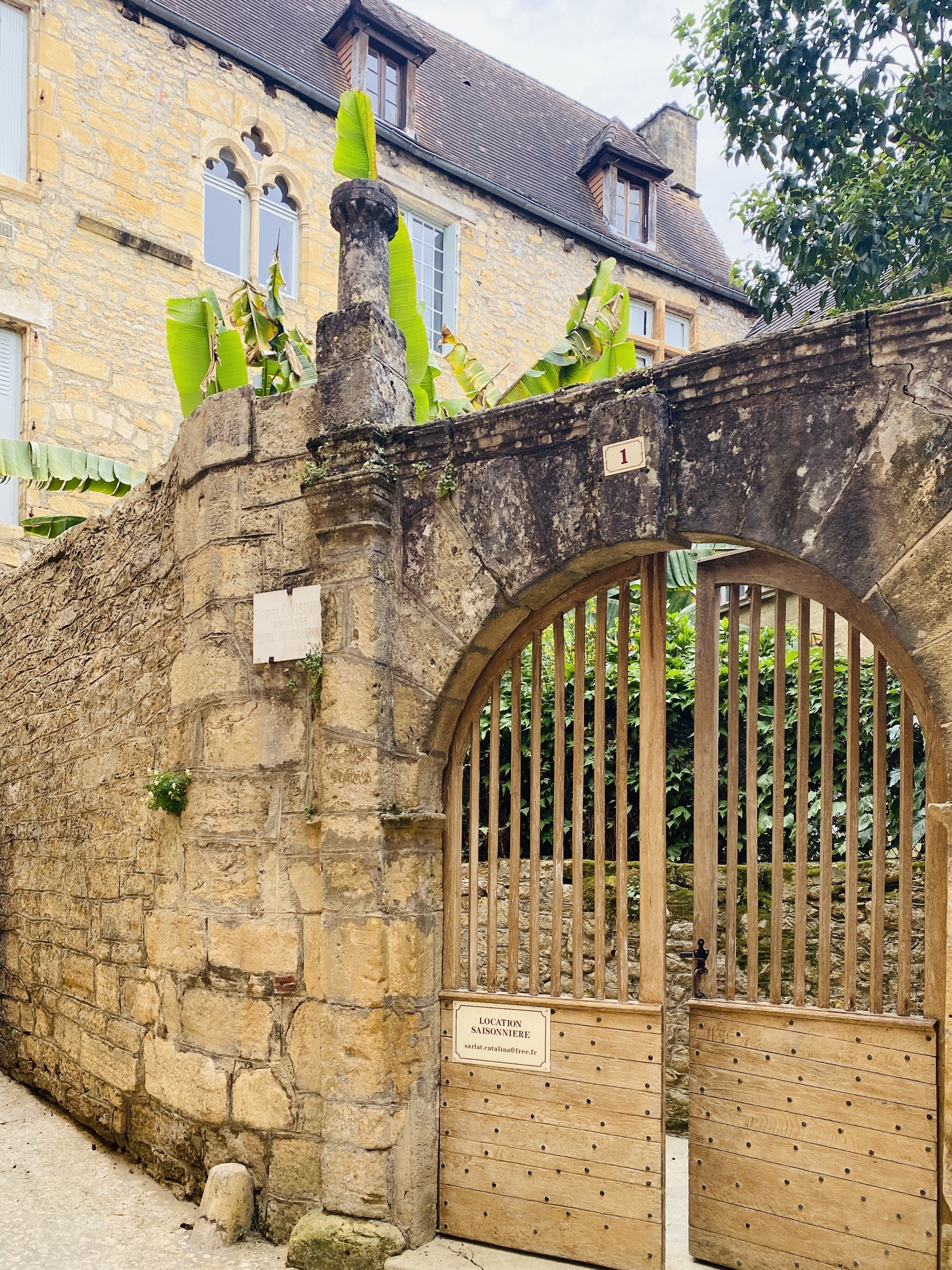 A stone wall with a wooden gate, a sign indicating 'Location Saisonnière', and a building with windows in the background.
