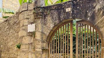 A stone wall with a wooden gate, a sign indicating 'Location Saisonnière', and a building with windows in the background.