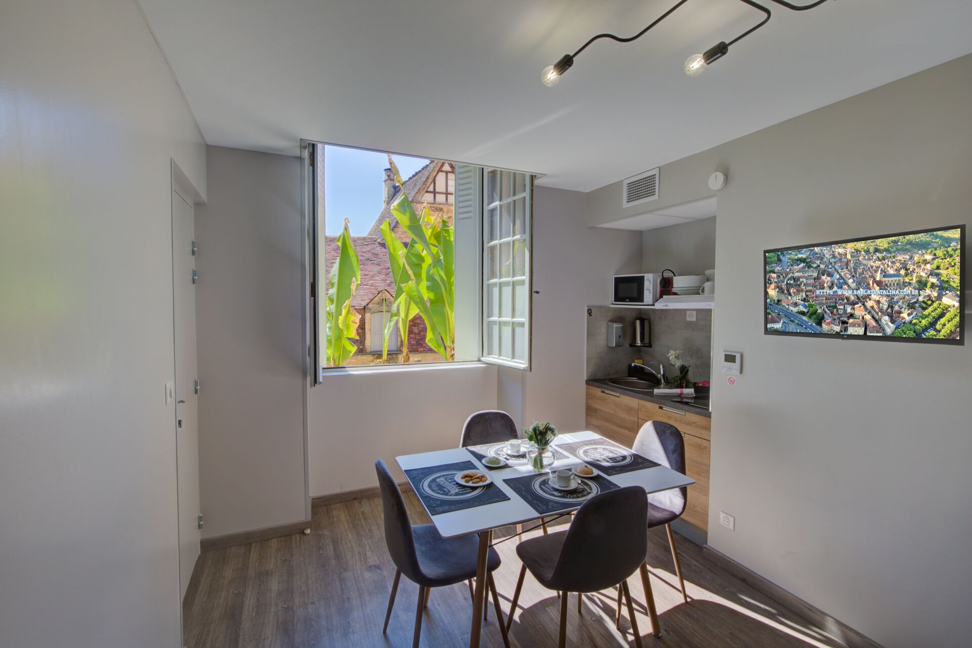 Un espacio habitable pequeño con zona de comedor, kitchenette y un amplio ventanal con vistas a un edificio y vegetación.