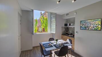 Un espacio habitable pequeño con zona de comedor, kitchenette y un amplio ventanal con vistas a un edificio y vegetación.