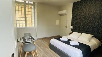 Et hotelværelse med en seng, et skrivebord og en stol. Der er et vindue med persienner, en aircondition og en tapetvæg.