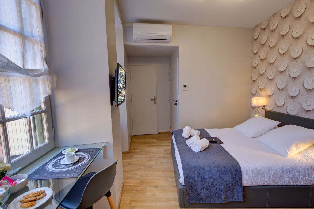 A hotel room with a bed, a desk with a chair, a window with curtains, and a wall with a circular pattern.