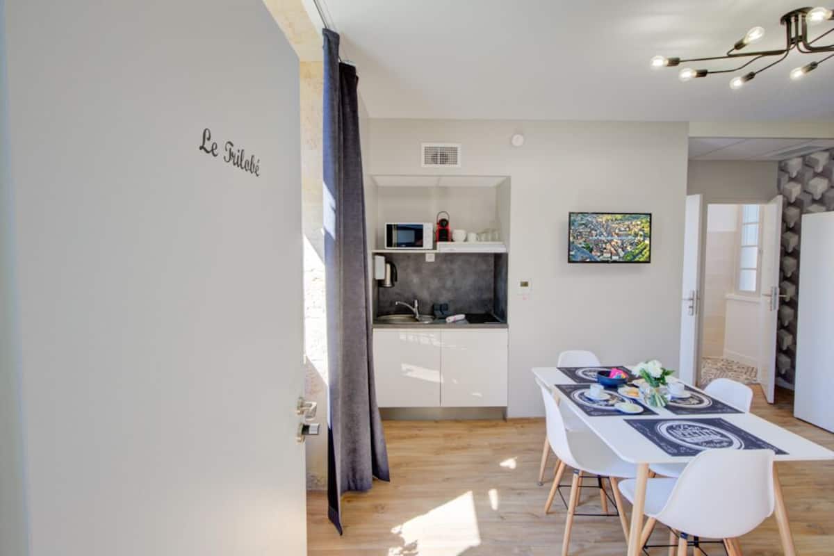 Een moderne keuken met witte kasten, een magnetron en een spoelbak. Er is een eethoek met een tafel gedekt voor twee personen, witte stoelen en een decoratieve vaas.
