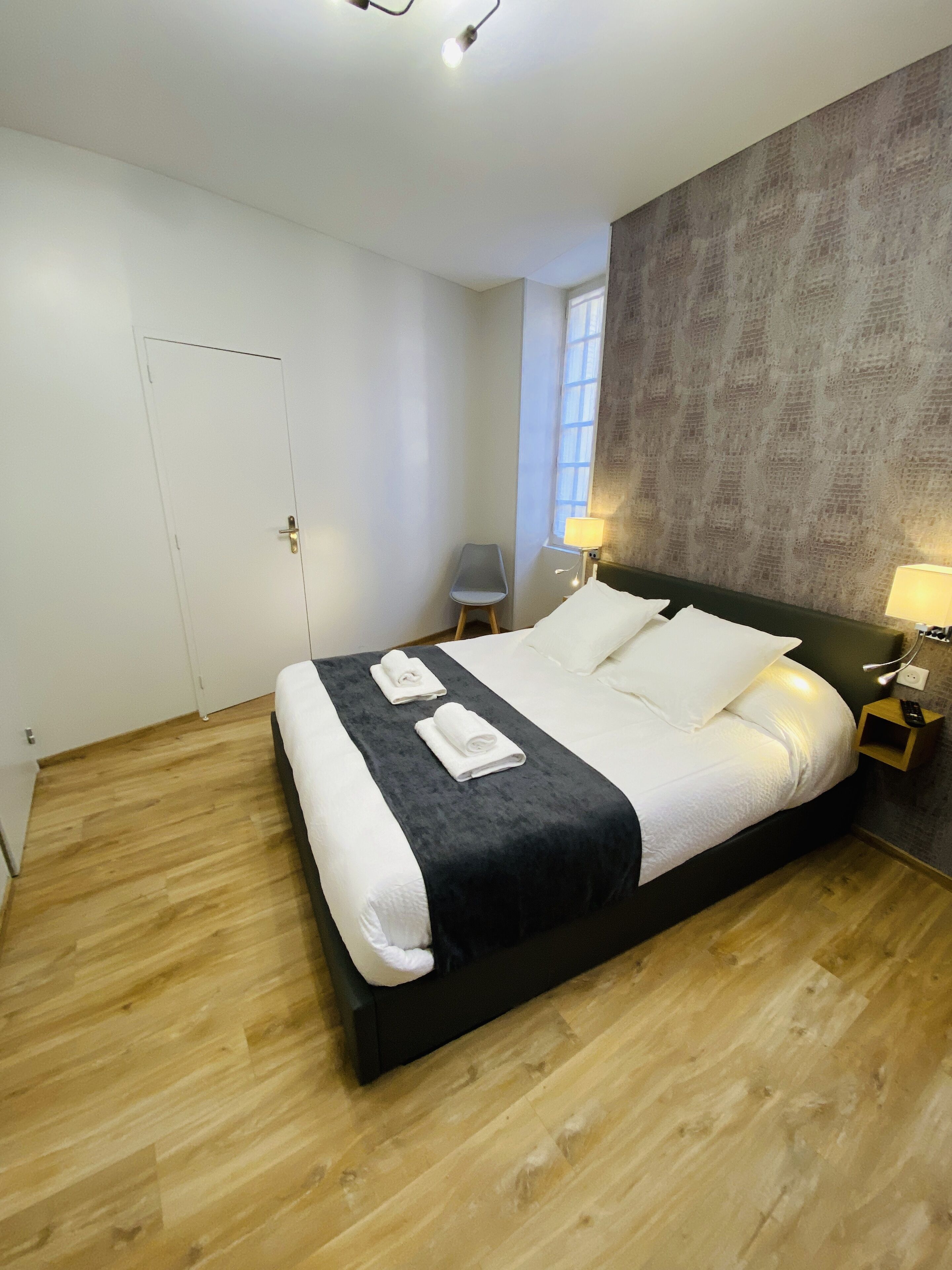 A modern bedroom with a large bed, a small chair, and a nightstand with a lamp.