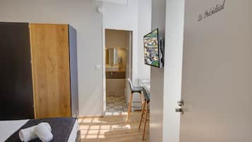 A bedroom with a bed, a wooden wardrobe, a desk with a chair, and a bathroom visible through an open door.