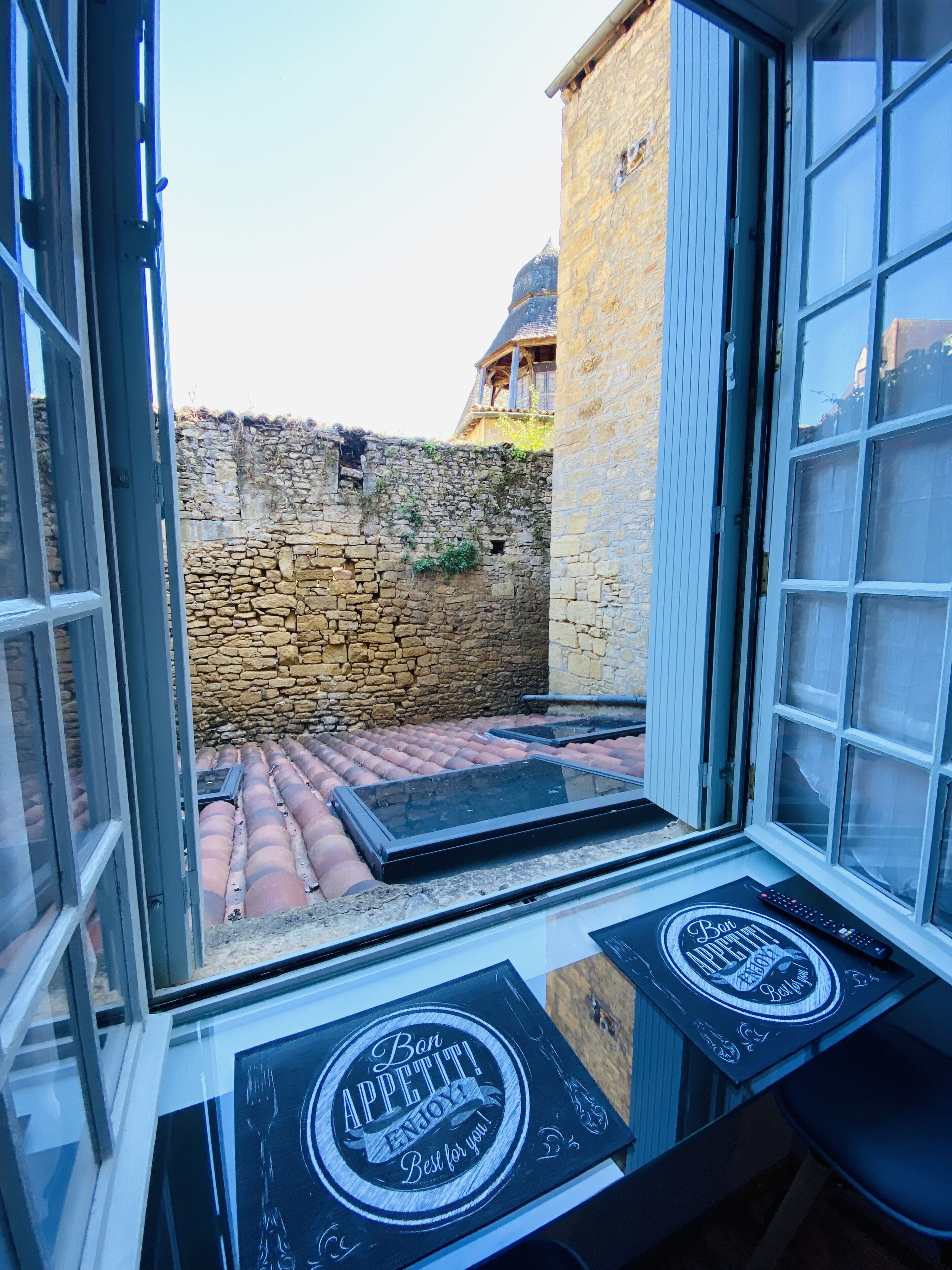 Una habitación con pared de piedra, una ventana que da a una pared de piedra y un edificio, y una mesa con mantel individual que dice "Bon APPETIT! ENJOY Best for you!".
