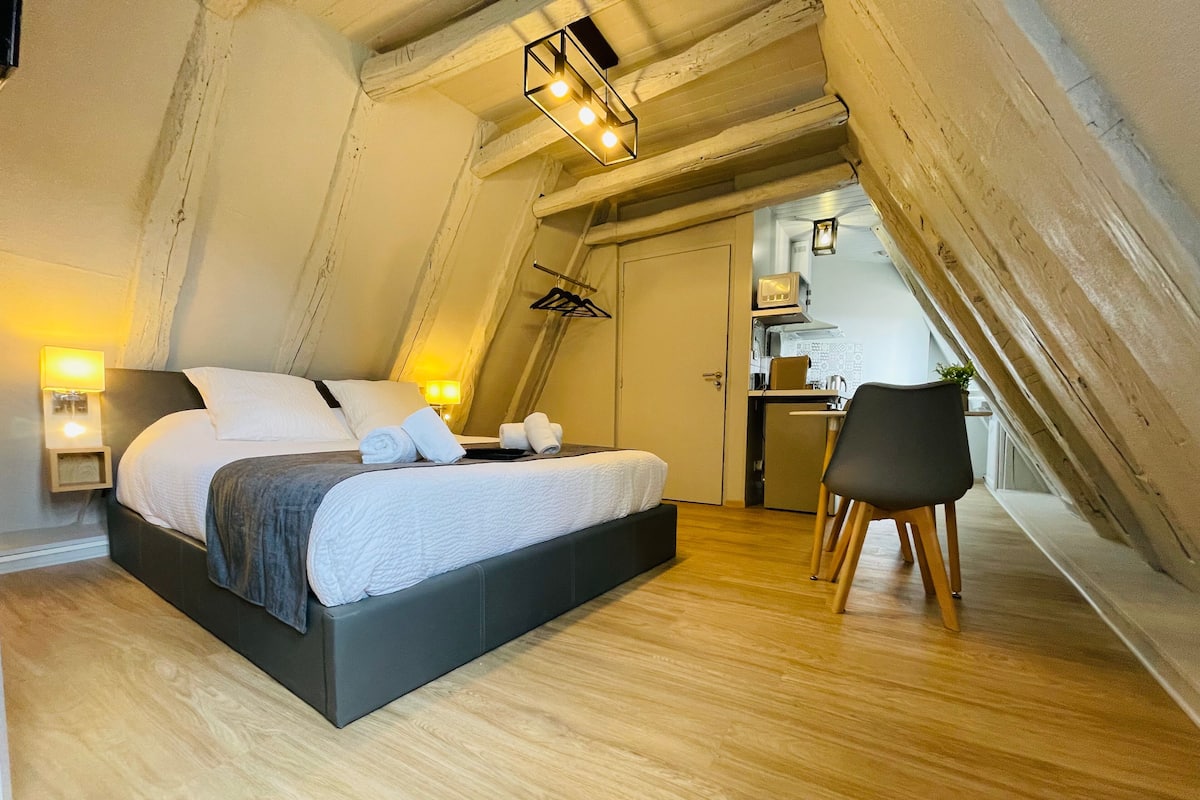 Een slaapkamer met een bed, nachtkastjes, een stoel en een kitchenette op de achtergrond.