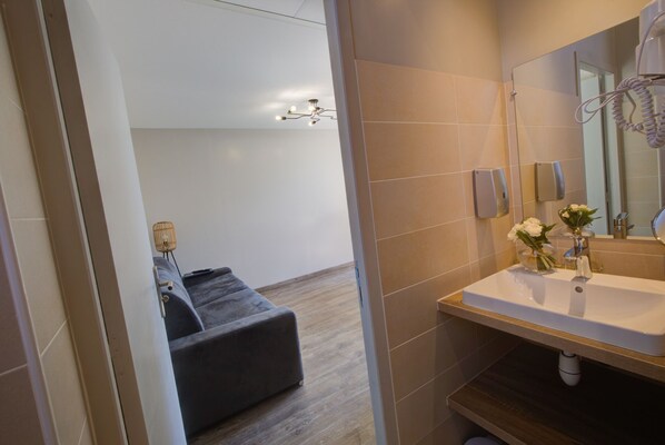 Un baño moderno con un espejo grande, un lavamanos blanco y un sofá gris al fondo.