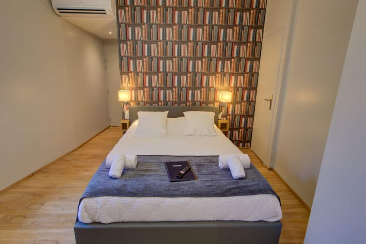 Een opgemaakt bed met witte lakens en een grijze deken, met aan weerszijden nachtlampjes, in een kamer met een wand vol boeken en een houten vloer.