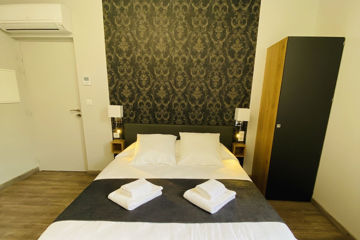 Een netjes opgemaakt bed met witte lakens, een donker hoofdbord met patroon en twee bedlampjes.
