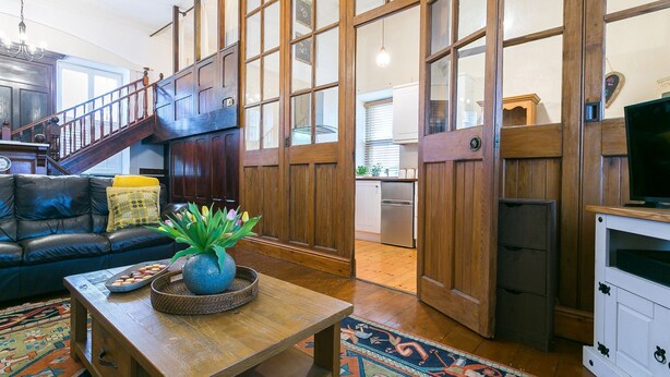 A spacious living area with a dark wooden staircase, a leather sofa, a coffee table with a vase, and a kitchen in the background. - Capel Bach  -  a near a pub that sleeps 6 guests  in 3 bedrooms (Llanystumdwy)