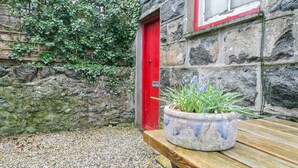 A stone building with a red door, a wooden bench, and a potted plant with blue flowers. - Capel Bach  -  a near a pub that sleeps 6 guests  in 3 bedrooms (Llanystumdwy)