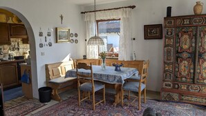 Un coin repas avec une table dressée pour quatre personnes, entourée de chaises en bois et d’un banc. Un tapis à motifs recouvre le sol, et un meuble décoratif se trouve à droite.